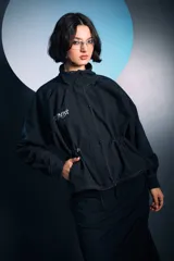 Campera negra de cuello alto con cierre frontal de botones a presión, mangas largas y cordones ajustables en la cintura. Presenta costuras a la vista en color blanco y un bordado decorativo en el pecho.