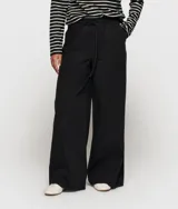 Pantalón ancho negro de corte relajado, confeccionado en mezcla de algodón y lino. Presenta cinturilla elástica de punto con cordón y trabillas, bolsillos laterales inclinados, etiqueta sobre el bolsillo izquierdo, minirayas bordadas bajo el bolsillo izquierdo y un bolsillo de ribete doble en la espalda.