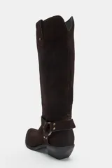 Bota alta de serraje marrón oscuro, de estilo texano, con tacón en bloque y puntera ligeramente puntiaguda. Cuenta con detalle de tiras cruzadas con tachas metálicas y una arandela en el tobillo, además de trabillas laterales en la parte superior de la caña.