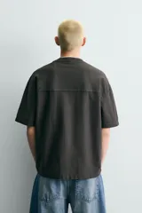 Camiseta relaxed fit de color gris con efecto lavado, cuello en pico y manga corta. Cuenta con múltiples estampados contrastantes en el frente, incluyendo texto y diseño heráldico.