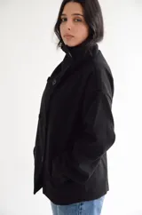 Abrigo negro de paño con diseño cruzado, cuello alto y cierre frontal con botones. Presenta una silueta estructurada y bolsillos laterales.