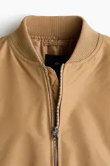 Campera bomber de nylon color tostado, con cuello, puños y bajo acanalados. Presenta cierre frontal, bolsillos laterales con botón a presión y un bolsillo con cierre en la manga.