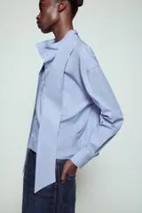 Camisa de algodón color celeste, con cuello alto y lazo integrado en el mismo tejido. Presenta cierre frontal con botones, manga larga con puños y un bolsillo frontal en el pecho. Diseño de corte recto.