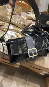 Cartera tipo bandolera de cuero charolado negro, con solapa frontal, tira central con hebilla metálica grande y correa regulable.