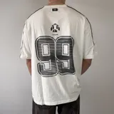 Remera oversize blanca de manga corta con detalles de líneas negras en las costuras de los hombros y mangas. Presenta un gran estampado en la espalda con el número 99 en diseño rayado y el logo de la marca en la parte superior.