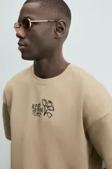 Sudadera de manga corta color beige, con cuello redondo y corte relajado. Presenta un bordado y estampado en el pecho con texto y un diseño floral.