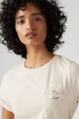 Remera color crudo de corte recto, con cuello redondo y mangas cortas con dobladillo. Tiene un bolsillo en el pecho con bordado de una flor.