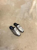 Suecos tipo mocasín de cuero metalizado plateado, con detalle de estribo metálico en el empeine y suela plana negra.