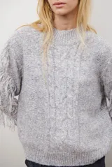 Buzo de punto color beige con cuello redondo, mangas largas con flecos decorativos y diseño de trenzas en el frente.