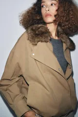 Chaqueta tipo bomber de color beige, con cuello solapa y aplicación extraíble de pelo sintético. Presenta cierre frontal con botones a presión ocultos, bolsillos delanteros y bajo con elástico.