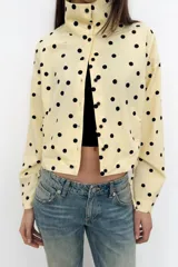 Chaqueta de cuello subido y manga larga con estampado de lunares negros sobre base beige. Cuenta con bolsillos de vivo en los laterales, detalles de frunces ajustables con cordones en el delantero y cierre frontal con botones.