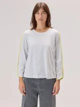 Remera de manga larga color gris claro, con detalle de franjas laterales en contraste color amarillo en las mangas. Presenta un diseño holgado y moderno.