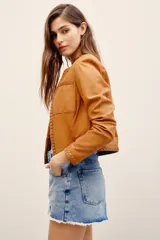 Saco corto de cuero ovino en color tostado, sin cuello, con detalles de trenzados en los bordes y bolsillos frontales con solapa.
