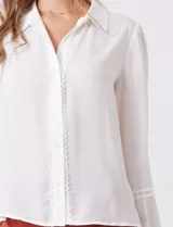 Camisa de gasa blanca con cuello solapa, cierre frontal con botones y detalle de puntilla a lo largo de la abertura. Presenta mangas largas con puños elásticos y costuras decorativas en el antebrazo.