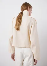Chaqueta crop de lino color beige, diseño recto, cuello alto tipo embudo, manga larga, dos bolsillos laterales y cierre cruzado de doble botonadura metálica dorada con detalles en los hombros.