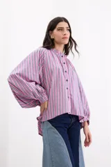 Camisa de corte holgado con diseño de rayas verticales en tonos fucsia, blanco y azul. Presenta cuello redondo con volado, cierre frontal con botones azules y mangas largas abullonadas con hombros caídos.