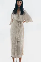 Vestido midi de Zara Woman Collection color beige, con cuello solapa, mangas por debajo del codo y diseño cruzado. Confeccionado en tejido plisado con brillo satinado, incluye un cinturón ajustable del mismo género con lazada en la cintura.