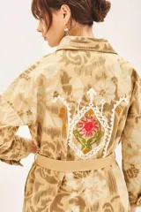 Campera de corte recto confeccionada en gabardina de algodón, con diseño estampado y detalles bordados. Presenta cuello camisero, cierre frontal con botones, bolsillos con solapa en el pecho y lazo ajustable en la cintura con terminaciones bordadas.