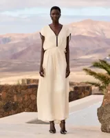 Vestido largo color crudo, confeccionado en viscosa satinada de inspiración grecorromana. Cuenta con escote en V profundo con ribete negro en contraste, mangas cortas fluidas con detalles oscuros, y corte en la cintura con drapeado suave.
