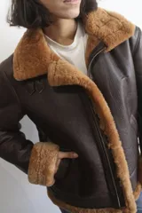 Campera bomber de cuero genuino color marrón oscuro, con interior y detalles en piel sintética color crema. Presenta cierre frontal, cuello solapa amplio y correas ajustables en el cuello y la cintura.