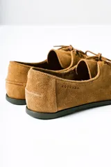 Mocasín de estilo relajado confeccionado en cuero gamuzado color tostado. Presenta detalles fruncidos en la puntera, cordones decorativos en el empeine y el logo de la marca grabado en el lateral.