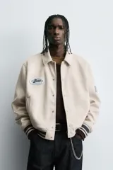 Cazadora estilo varsity de tejido efecto ante en color beige. Presenta cuello solapa, cierre frontal con botones a presión y puños y ruedo con acabado en rib a rayas negras. Incluye parches bordados decorativos en el pecho y la manga, además de bolsillos laterales de doble vivo.
