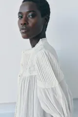 Camisa blanca de algodón con cuello redondo y manga por debajo del codo abullonada. Presenta detalles de bordados a tono y cierre frontal con botones.