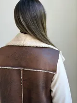 Chaleco de cuero sintético color marrón chocolate con interior y solapas de corderito sintético color crudo. Diseño sin mangas con cierre frontal.