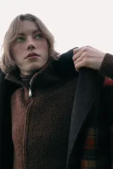 Capa oversize de corte amplio con estampado de cuadros en tonos rojos, negros y marrones. Presenta cuello alto, cierre frontal con botones, bolsillos delanteros y mangas ajustables mediante trabillas con botones.