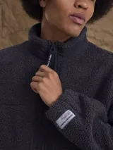 Campera de sherpa color verde oliva con cierre frontal completo, cuello alto y detalles en contraste negro en la cremallera. Presenta una etiqueta decorativa en el puño.