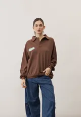 Buzo celeste de manga larga con cuello polo y cierre frontal de botones. Presenta un parche marrón bordado en el pecho.