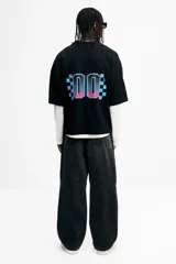 Remera de malla negra con cuello en V, mangas cortas y hombros caídos. Presenta un estampado frontal con la frase "THUNDER X RACING TEAM" y un diseño gráfico en tonos rosa, azul y celeste con la palabra "RACING" y un patrón de cuadros.