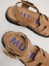 Sandalias de cuero marrón con tiras entrecruzadas, suela de goma negra con pespuntes blancos y hebilla lateral.