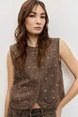 Chaleco de denim marrón con efecto acid wash y apliques de strass distribuidos por toda la prenda, sin mangas y con cierre frontal de botones.