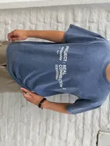 Remera oversize de algodón con efecto acid wash en tono azul oscuro, con estampa blanca en la espalda que incluye los textos "Ost" y "Osten" enmarcados en óvalos, y un diseño de globo terráqueo en el centro.