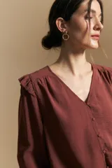 Camisa de manga larga color bordó, con escote en V y cierre frontal de botones forrados. Presenta detalles de volados en los hombros y frunces que aportan movimiento a la prenda.