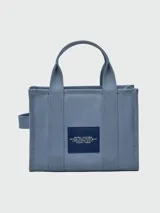 Bolso tipo tote de lona color beige de la marca Marc Jacobs. Presenta el texto 'THE TOTE BAG' estampado en el frente en color blanco, dos asas de mano superiores y una correa de hombro ajustable y removible en color negro.