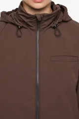 Gabardina de largo medio con corte en línea A y diseño holgado, confeccionada en tejido técnico color marrón. Cuenta con cierre frontal con cremallera, cuello alto con capucha integrada, mangas largas ajustables con trabilla y bolsillos laterales.