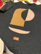 Campera de paño negro con diseño de collage abstracto en el frente, compuesto por parches de tela texturizada en tonos mostaza y rosado. Presenta una etiqueta de cuero sintético con el logo de la marca en la parte inferior.