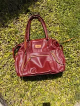 Bolso maternal de cuero marrón con doble asa de hombro, correa ajustable, múltiples bolsillos frontales y laterales con cierre, y detalles de tiras trenzadas.