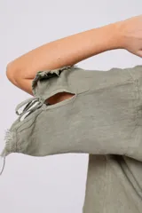 Blusa verde oliva de lino con cuello redondo, mangas cortas amplias con flecos, cierre lateral con botones y ruedo con flecos.