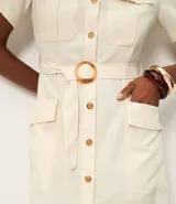 Vestido midi color crema, tipo camisero, con cuello, manga corta y cierre frontal de botones. Presenta bolsillos utilitarios en el pecho y la cadera, y un cinturón ajustable con hebilla redonda de madera. Confeccionado en sarga de algodón.