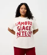 Remera de algodón de corte holgado y largo, con cuello redondo y mangas cortas. Presenta una estampa frontal con la frase 'L'AMORE GIACE IN TE' en color rojo sobre fondo crema.