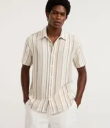 Camisa de hombre de manga corta con cuello tradicional y abotonado frontal. Presenta un tejido texturizado con un patrón de rayas verticales en tonos beige, blanco y marrón claro.