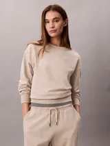 Buzo de felpa Calvin Klein de color beige, con cuello redondo, manga larga y detalles de canalé en cuello, puños y bajo. Presenta un emblema con logo texturizado en el pecho.