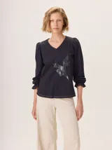 Remera de manga larga color negro con cuello en V y puños fruncidos. Presenta un estampado sutil en tonos grises y blancos en el centro del pecho, simulando un ramillete de flores secas.