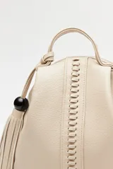 Mochila de símil cuero texturizado color beige, con asa superior, tiras regulables para la espalda y cierre principal metálico. Presenta un detalle trenzado vertical en el frente y un accesorio con flecos y cuenta negra en el lateral.
