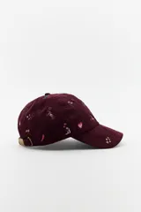 Gorra tipo baseball confeccionada en pana color bordó, con múltiples bordados decorativos de motivos variados como cactus, caballos, corazones y estrellas. Presenta ajuste posterior y visera curva.