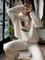Buzo tipo hoodie de corderito color manteca, con capucha y cordones ajustables. Presenta un diseño abstracto frontal con apliques de textura suave en tonos rosa viejo y marrón.