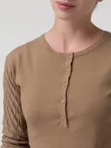 Remera de manga larga color beige, con cuello redondo y cierre frontal parcial con botones. Presenta un diseño de tejido trenzado con textura de rombos en las mangas y cuerpo liso.